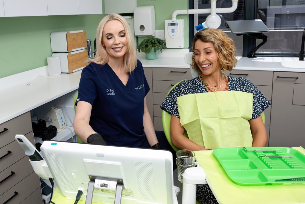 Dr. Sheryl Smithies demonstrating a digital 3D teeth model on a touchscreen monitor to a smiling patient.